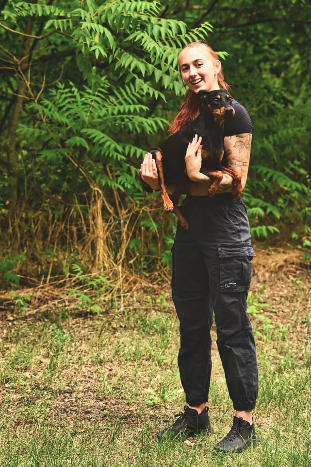 Lou steht im Farn und hält einen kleinen schwarzen Hund